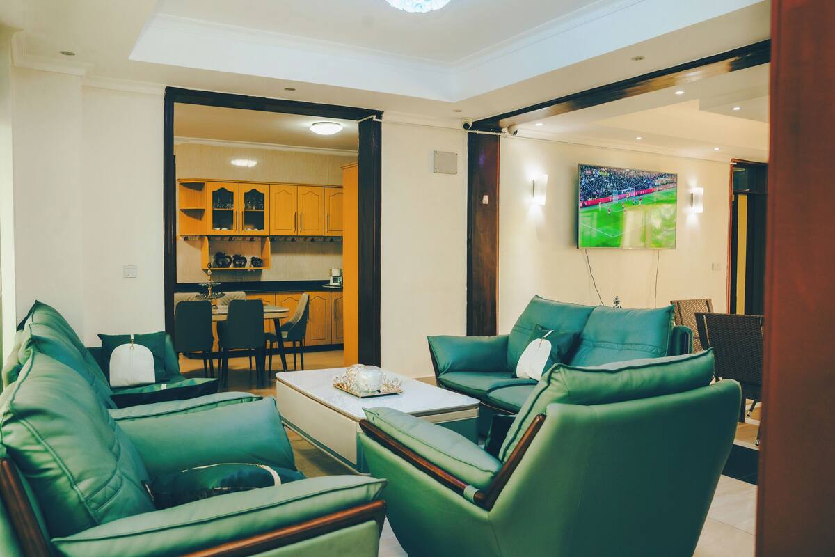 A modern living room with green leather sofas, a coffee table, and a kitchen in the background.