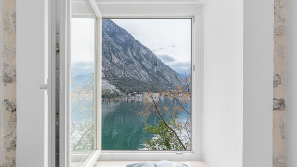 Une pièce avec une fenêtre donnant sur un lac et des montagnes, un livre posé sur le rebord de la fenêtre et un mur blanc.
