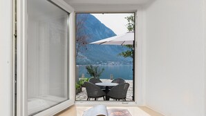 Une porte coulissante s’ouvre sur un balcon aménagé avec une table et des chaises, d’où l’on peut admirer un lac et des montagnes.