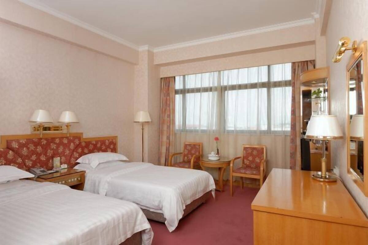 Habitación de hotel con dos camas, un escritorio, una silla, una mesita pequeña y un ventanal grande con cortinas.
