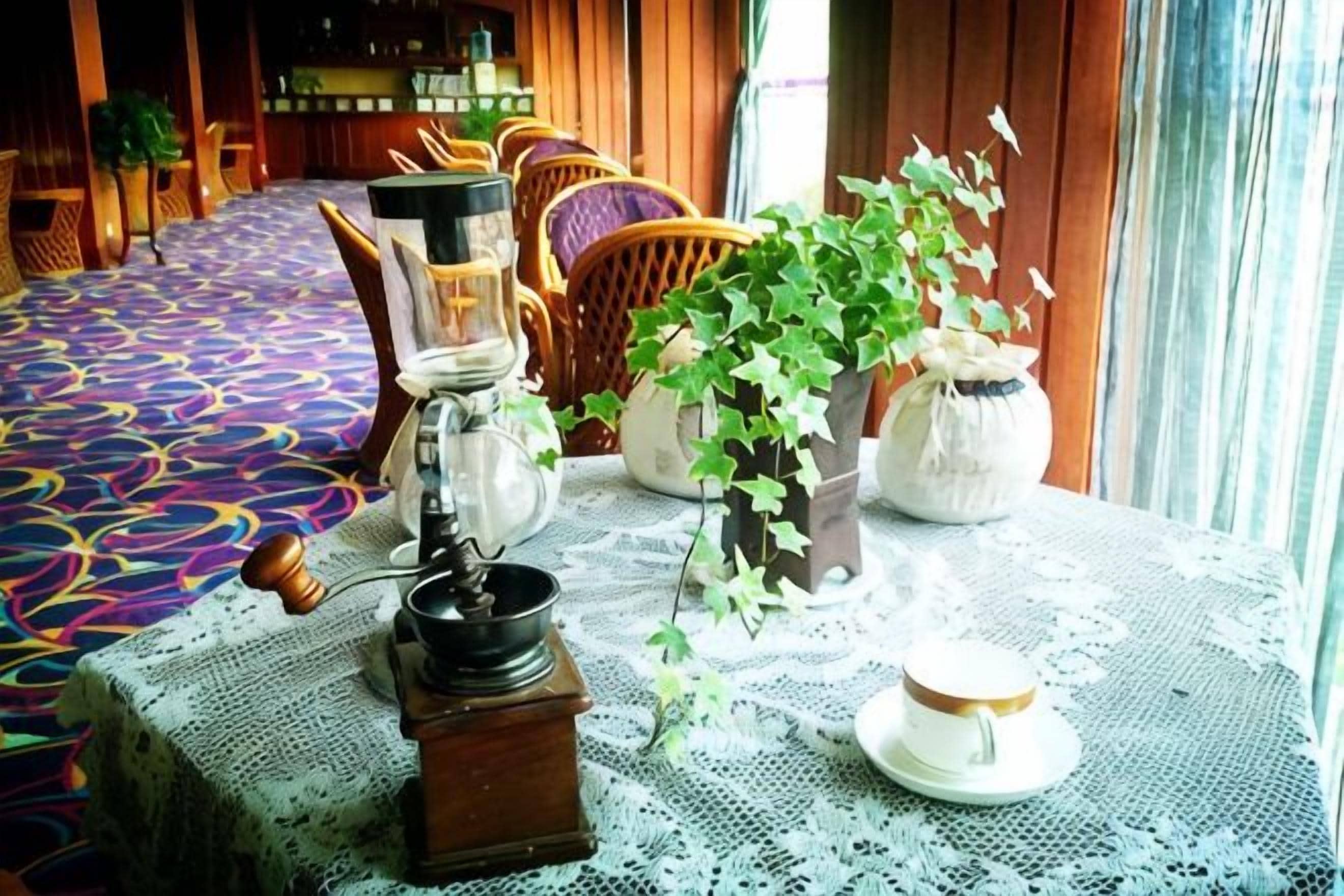 Una habitación con alfombra estampada, una mesa con mantel de encaje, un molinillo de café y plantas en macetas.