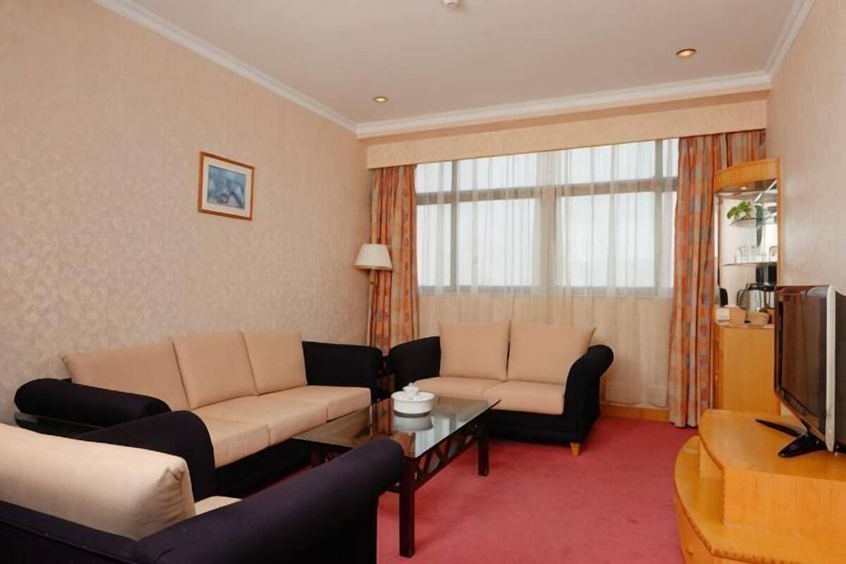 Habitación de hotel con sofá, mesa de centro, televisor y ventana con cortinas.