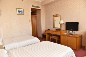 Ein Hotelzimmer mit zwei Betten, einem Schreibtisch mit Spiegel, einem Fernseher und einem kleinen Hocker.