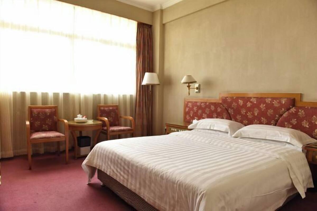 Habitación de hotel con una cama grande, dos sillas, una mesita, una lámpara y un ventanal con cortinas.