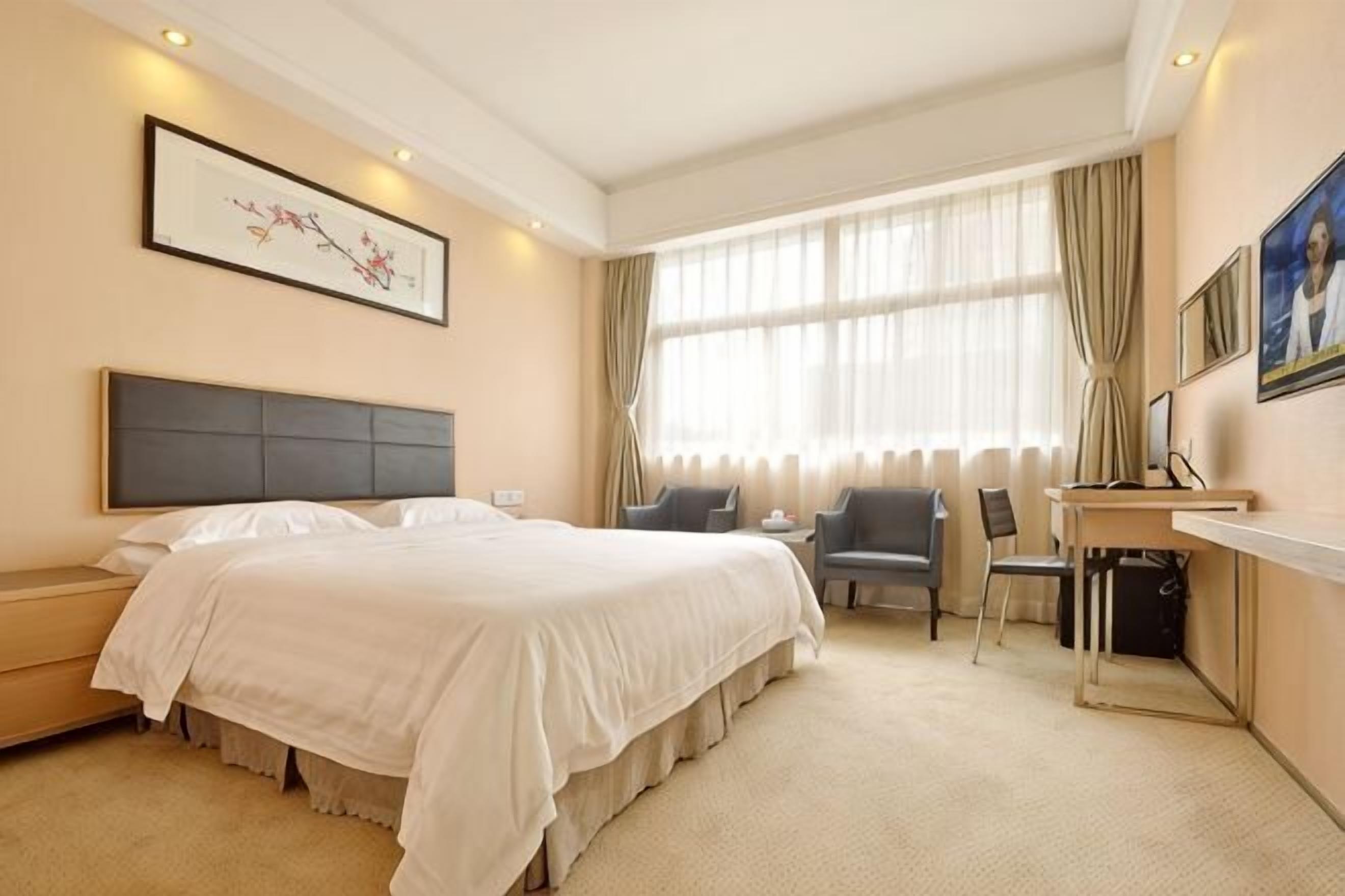 Hotellihuone, jossa on suuri sänky, työpöytä tuolilla, tietokone ja ikkuna, jossa on verhot.