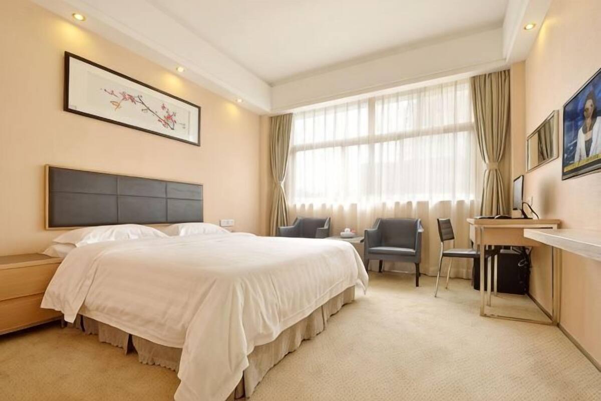 Habitación de hotel con una cama grande, un escritorio con silla, una computadora y un ventanal con cortinas.