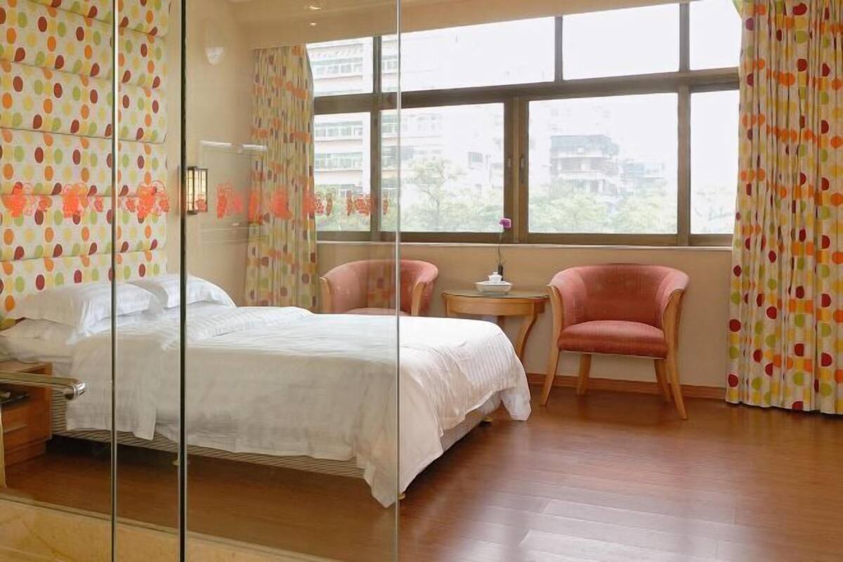 Un dormitorio con una cama, dos sillas, una mesita y un ventanal con cortinas.