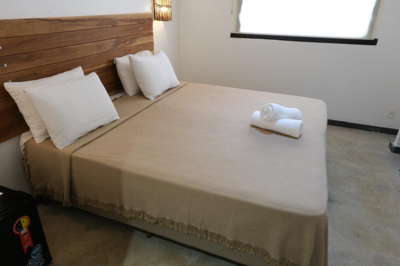 A neatly made bed with a beige bedspread and pillows, a wooden headboard, and a window with blinds.