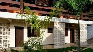 Un bâtiment moderne à la façade blanche, doté de balcons en bois et d’un jardin bien entretenu avec des palmiers et des pierres plates servant de chemin.