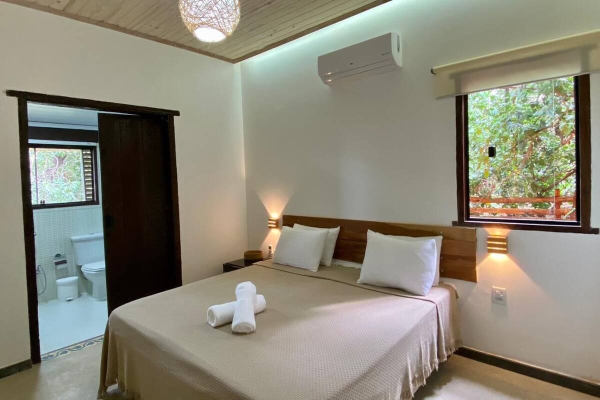 A bedroom with a bed, bedside lamps, a window with a view, and an air conditioning unit.