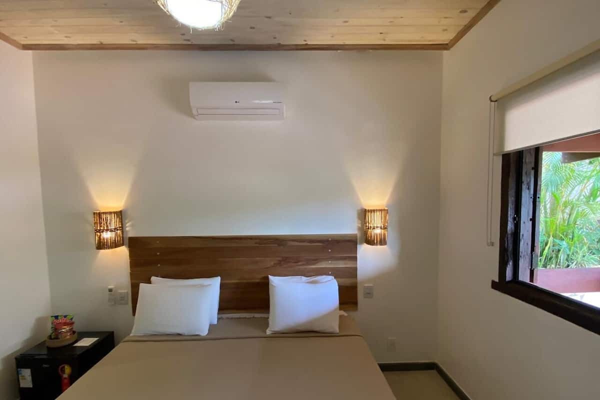 A hotel room with a wooden headboard, two white pillows, a bed, a nightstand with a lamp, and a window with a view of greenery.