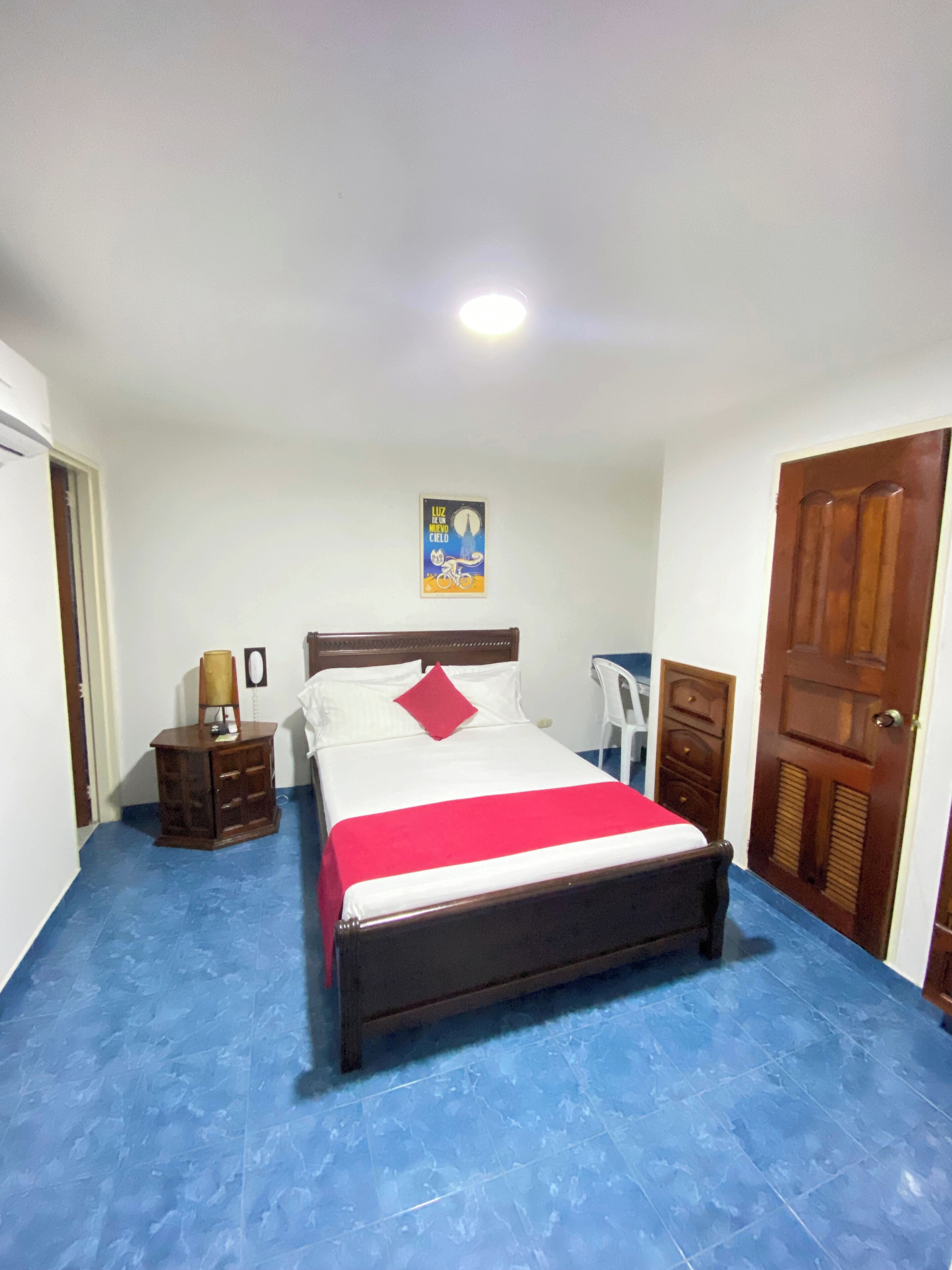 A bedroom with a bed, a nightstand, a chair, a wooden door, and a wall-mounted light fixture.