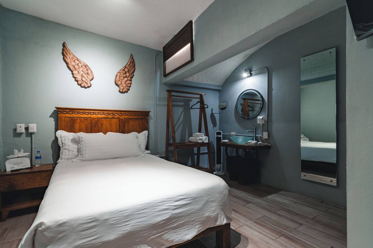 Een slaapkamer met een groot bed, een houten hoofdbord, een nachtkastje met een lamp, een spiegel, en een badkamer met een wastafel en spiegel.