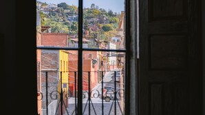 A view through a window with metal bars, featuring a cobblestone street, buildings, and a hillside in the background. - Casa Xaca (San Miguel de Allende)