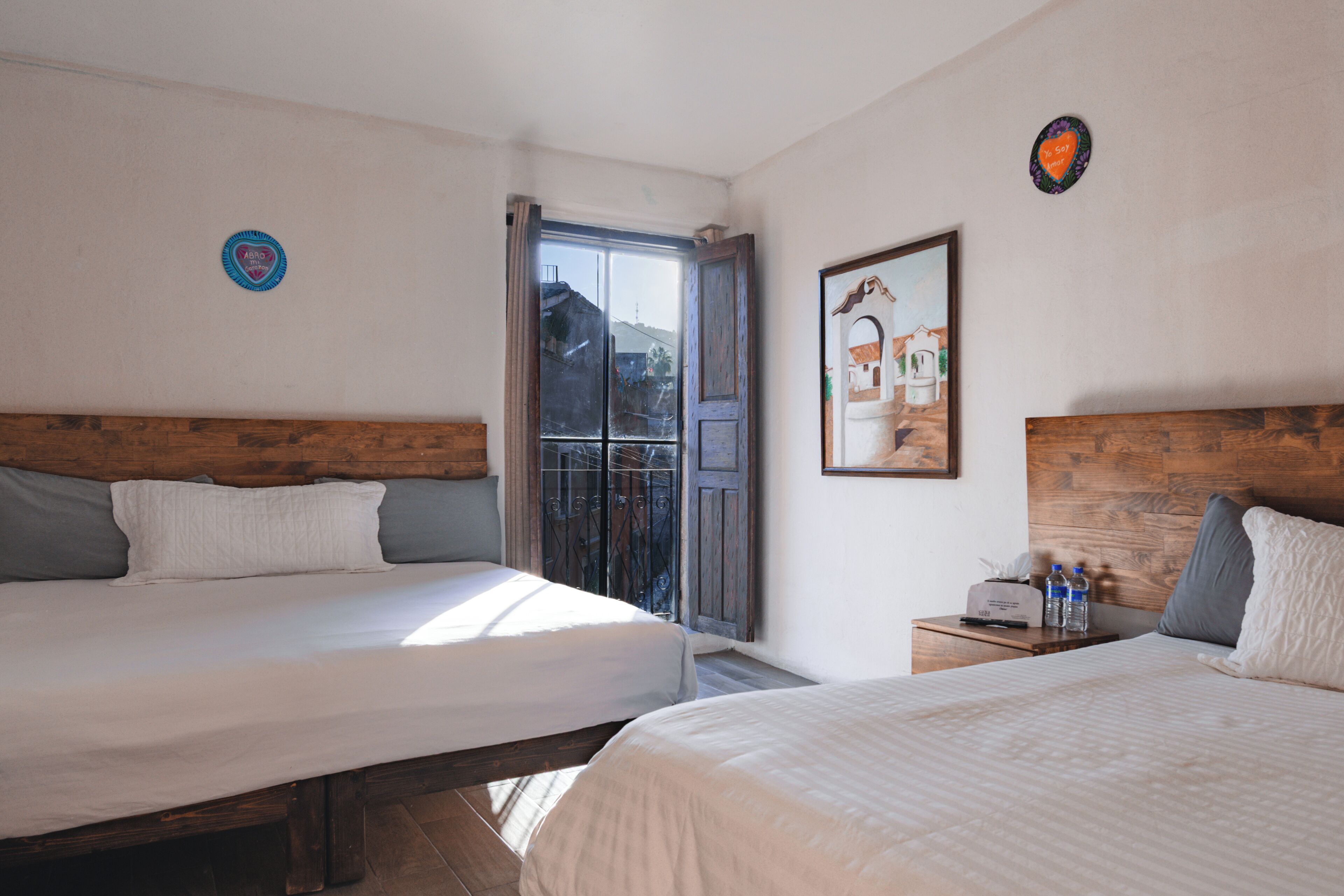 A hotel room with two beds, a wooden headboard, a nightstand with a clock and bottles, and a window with a view of buildings.