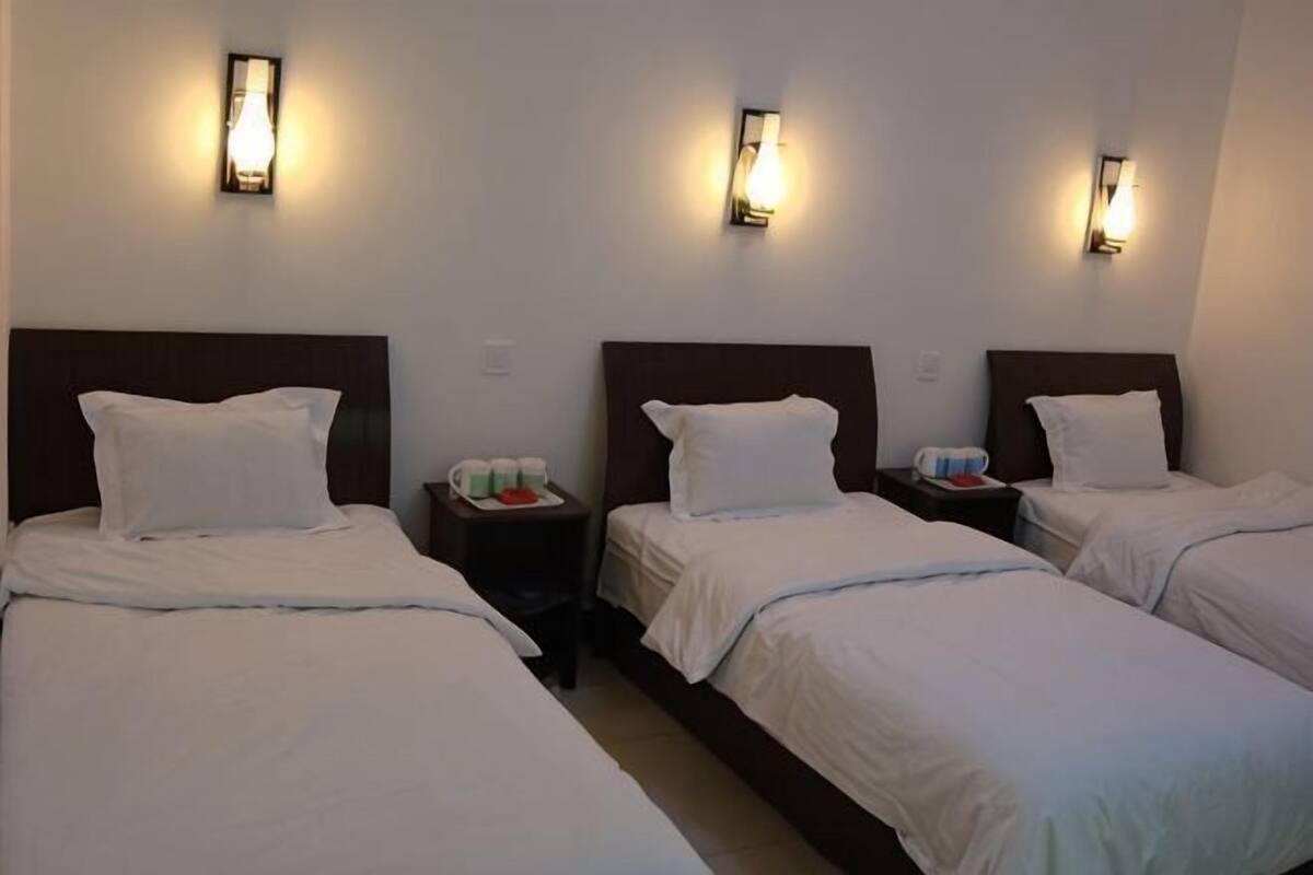 A hotel room with three single beds, each with a headboard and white bedding. There are bedside tables with lamps and a tray with cups and a red object.