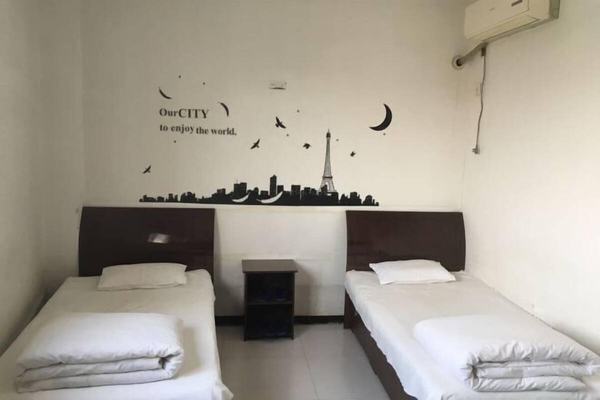 A hotel room with two single beds, a small nightstand, and a wall decal featuring a city skyline with a crescent moon and birds.
