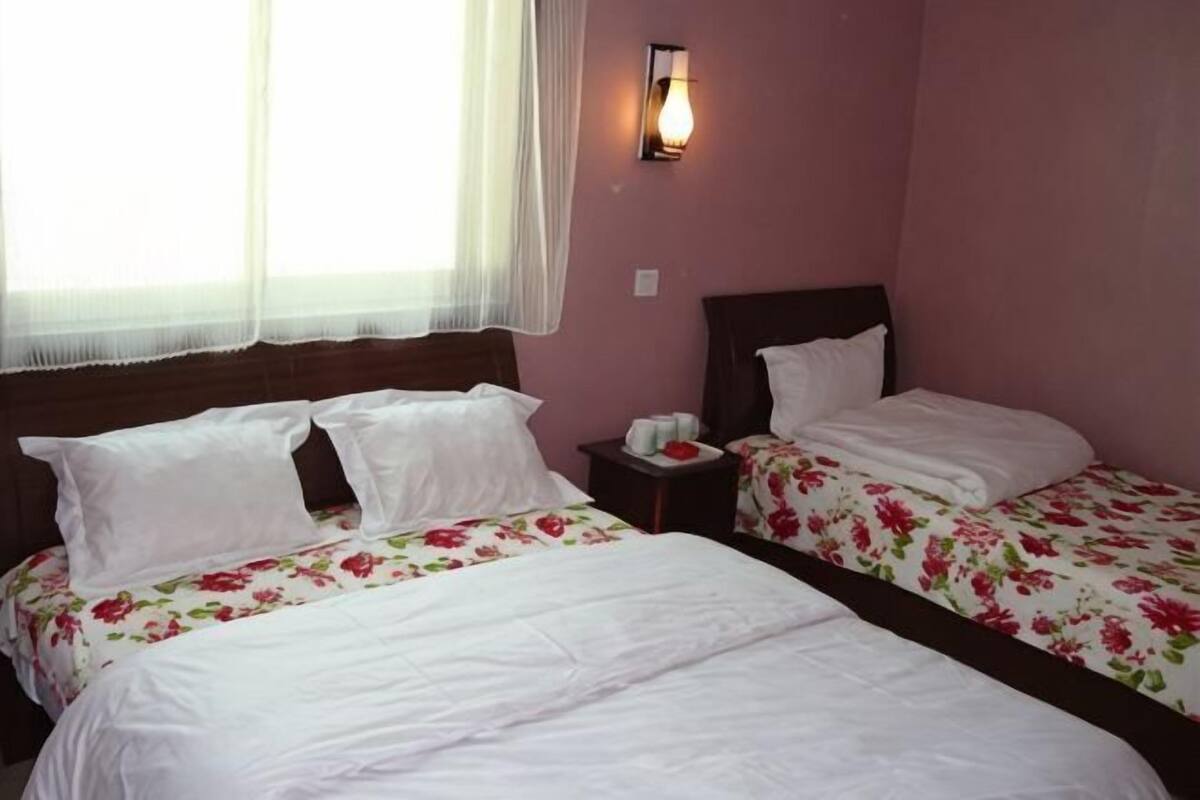 A room with two beds, a nightstand, a wall-mounted light, and a window with sheer curtains.