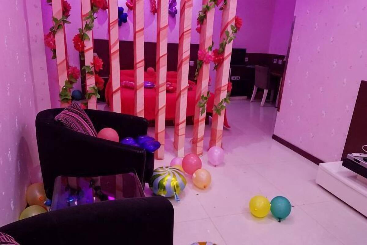 A room with a bed, a black chair, and a small table. There are balloons on the floor.