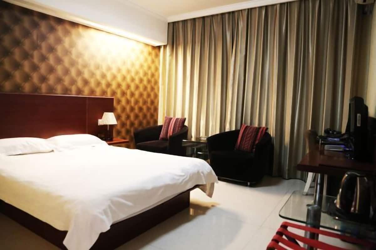 A hotel room with a large bed, a desk with a kettle, a chair, and a TV.