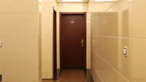 Um corredor estreito com uma porta marrom no final, paredes revestidas de azulejos e um interruptor de luz à direita.