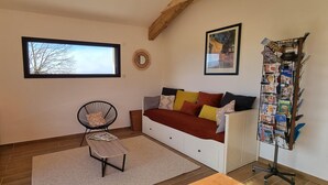 A cozy room with a wooden cabinet, a television, a chair, a sofa with colorful cushions, a small table, a window with a view of trees, a wall-mounted mirror, and a rack with magazines. - Gite facing the Vercors (Marches)