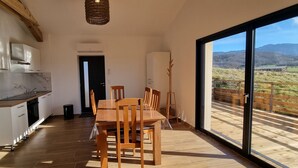 A modern kitchen with white cabinets and a marble countertop, a dining area with wooden chairs, and a large window with a view of a rural landscape. - Gite facing the Vercors (Marches)