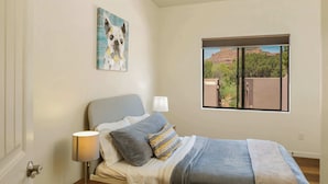 Ein Schlafzimmer mit Bett, Nachttisch, Lampe, einem Bild und einem Fenster mit Blick auf eine Berglandschaft.