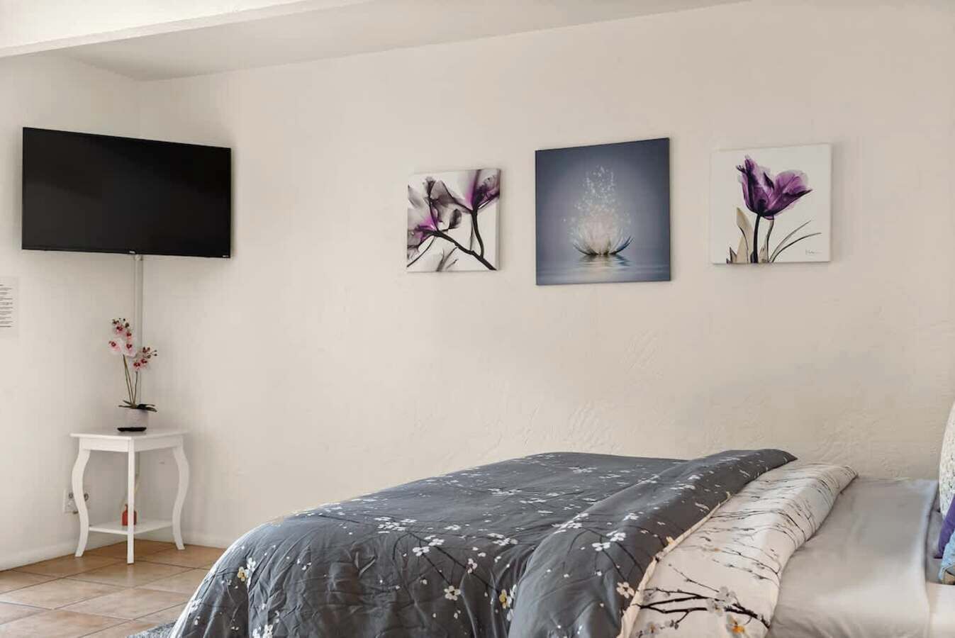 A bedroom with a bed, a TV mounted on the wall, and three framed floral artworks.
