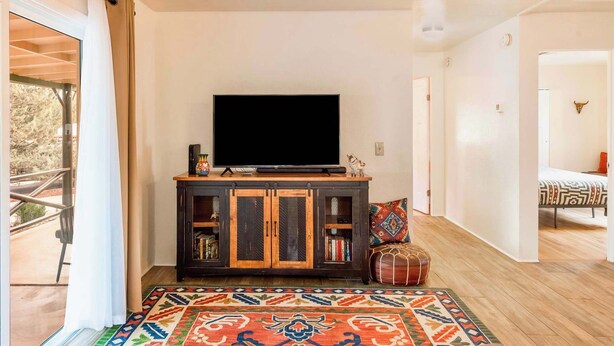 Een woonkamer met een televisie op een houten kast, een kleurrijk vloerkleed en een deur naar een andere kamer.