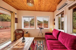 Une pièce avec un plafond en bois, un canapé rouge, une table sur laquelle est posée une lanterne, et deux fenêtres donnant sur un paysage rocheux.