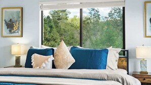 A bedroom with a large window, a bed with blue and beige bedding, two bedside tables with lamps, and framed artwork on the walls.