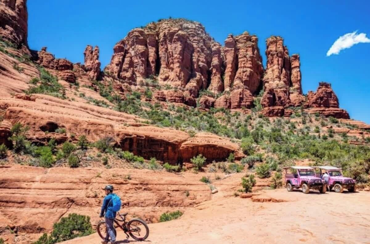 Un paysage désertique avec des formations rocheuses rouges, une personne debout à côté d’un vélo et un véhicule tout-terrain violet garé au sol.