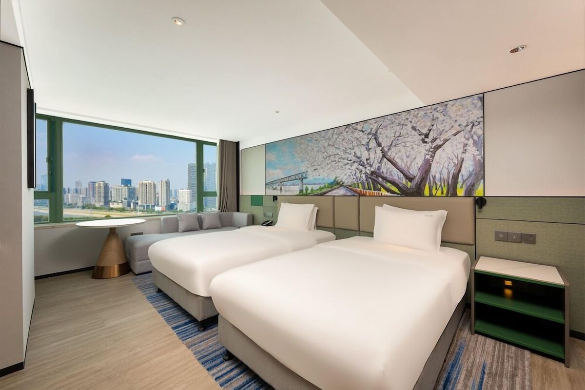 A hotel room with two beds, a view of the city, and a painting on the wall.