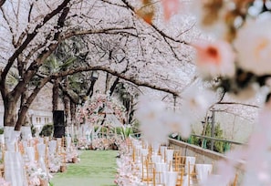 Un lieu de réception en plein air pour les mariages, avec une allée recouverte d’un tapis vert, des chaises blanches et des décorations florales.