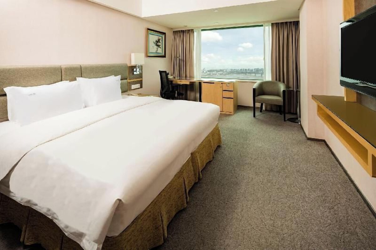 A hotel room with a large bed, a desk, a chair, a television, and a window with a city view.