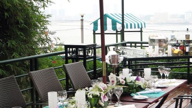 An outdoor dining area with wicker chairs, a table set with glassware, and a view of a building across the water. - Holiday Inn WUHAN RIVERSIDE by IHG (Wuhan)