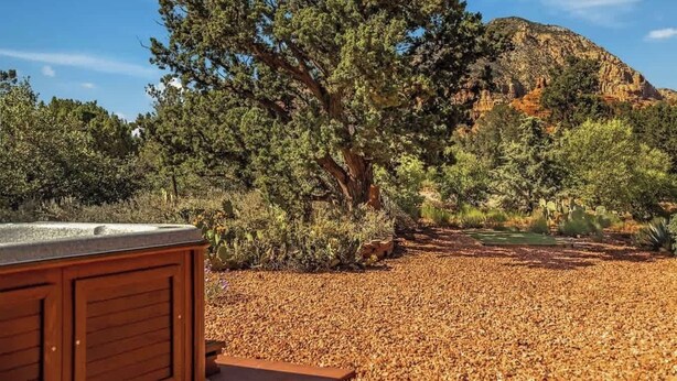 A wooden deck with a hot tub, surrounded by a dry, leaf-covered ground and a large tree. - Myrinn – Private Hot Tub & Red Rock Views (Sedona)