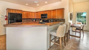 A modern kitchen with wooden cabinets, a large island with bar stools, and a dining area with a view. - Myrinn – Private Hot Tub & Red Rock Views (Sedona)