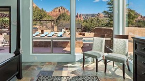 Habitación con ventanas amplias que ofrecen vista a una piscina y montañas. Hay dos sillas y una mesita en la habitación.
