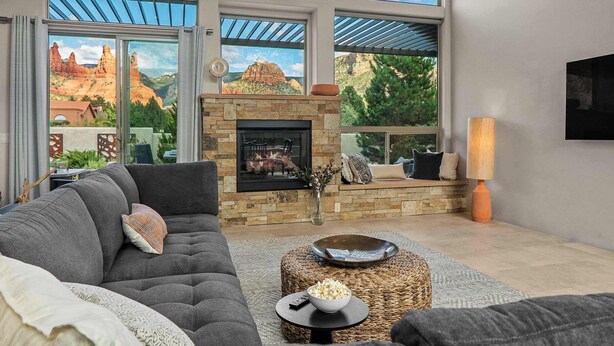 A modern living room with a stone fireplace, large windows with a scenic view, a comfortable sofa, and a coffee table with a bowl of popcorn. - Myrinn – Red Rocks Views & Cathedral Rock Trail (Sedona)