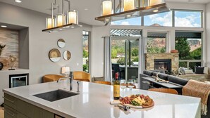 A modern kitchen with a large island, a dining area with a fireplace, and a view of the mountains. - Myrinn – Red Rocks Views & Cathedral Rock Trail (Sedona)