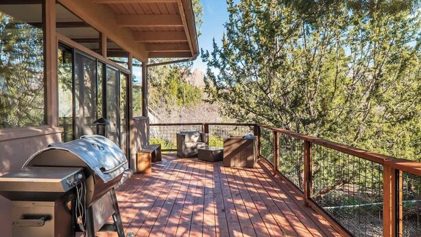Una terraza de madera con una parrilla, mobiliario exterior y vistas a árboles y un paisaje rocoso.