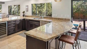 Una cocina moderna con una isla grande, armarios oscuros y vista a árboles a través de amplios ventanales.