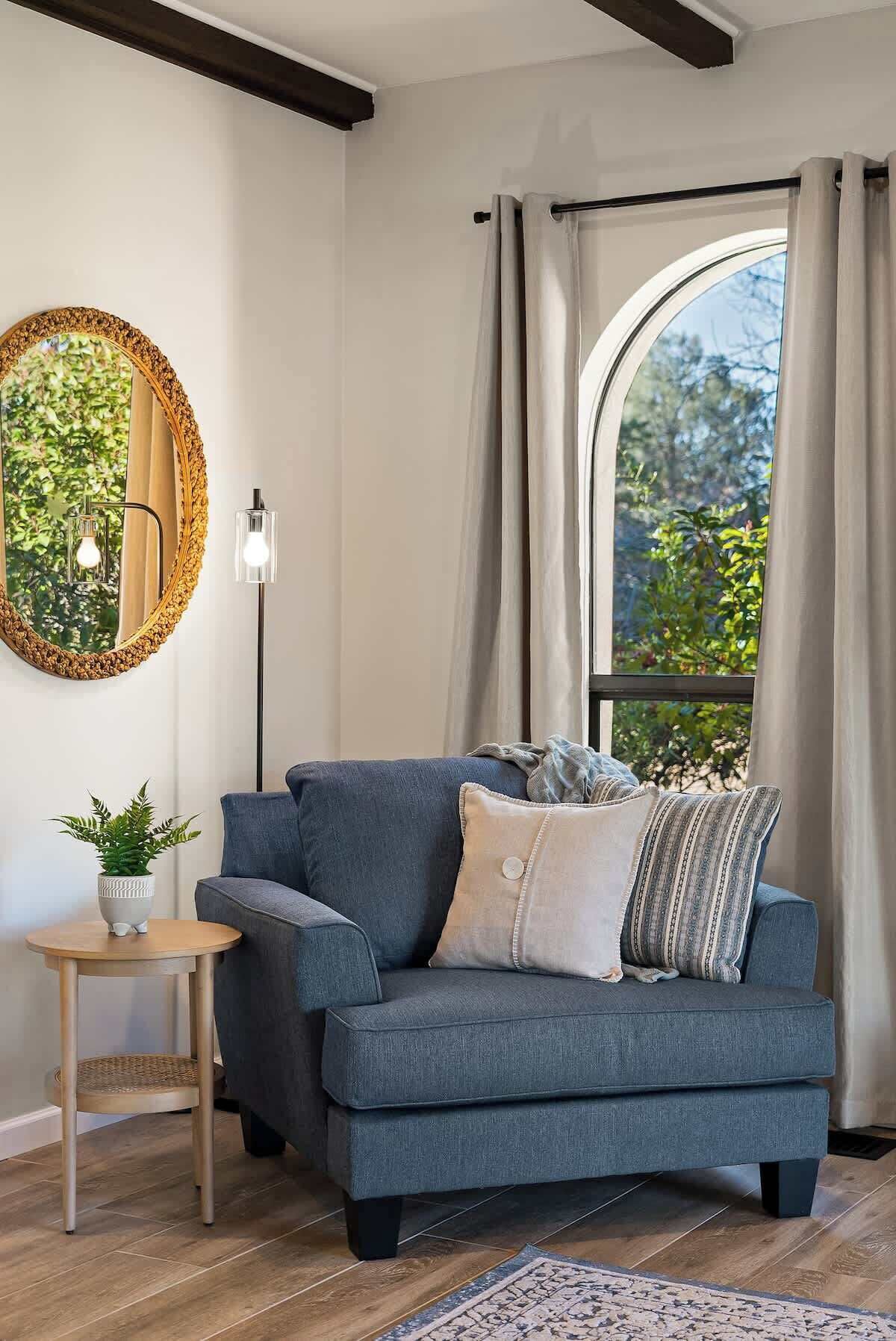 A modern living room with a grey sofa, a round mirror, a small side table with a plant, and a view through a large window with curtains.