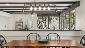Un comedor con una mesa de madera, sillas negras, un bol verde, una lámpara en el techo y una ventana con vistas a árboles.