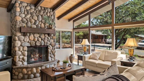 A stone fireplace, a TV, a sofa, and a dining table in a well-lit living room with large windows.