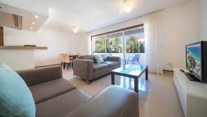 A modern living room with a sofa, a coffee table, and a television. There is a balcony with a view of palm trees. - Luxury 1 BR Condo with Pool - Kaan 304 (Playa del Carmen)