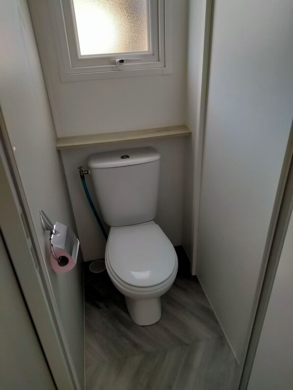 A small, narrow bathroom with a toilet, a window above it, and a wooden shelf. - 8-person mobile home, ideal for families! Sheets & towels included (Condrieu)
