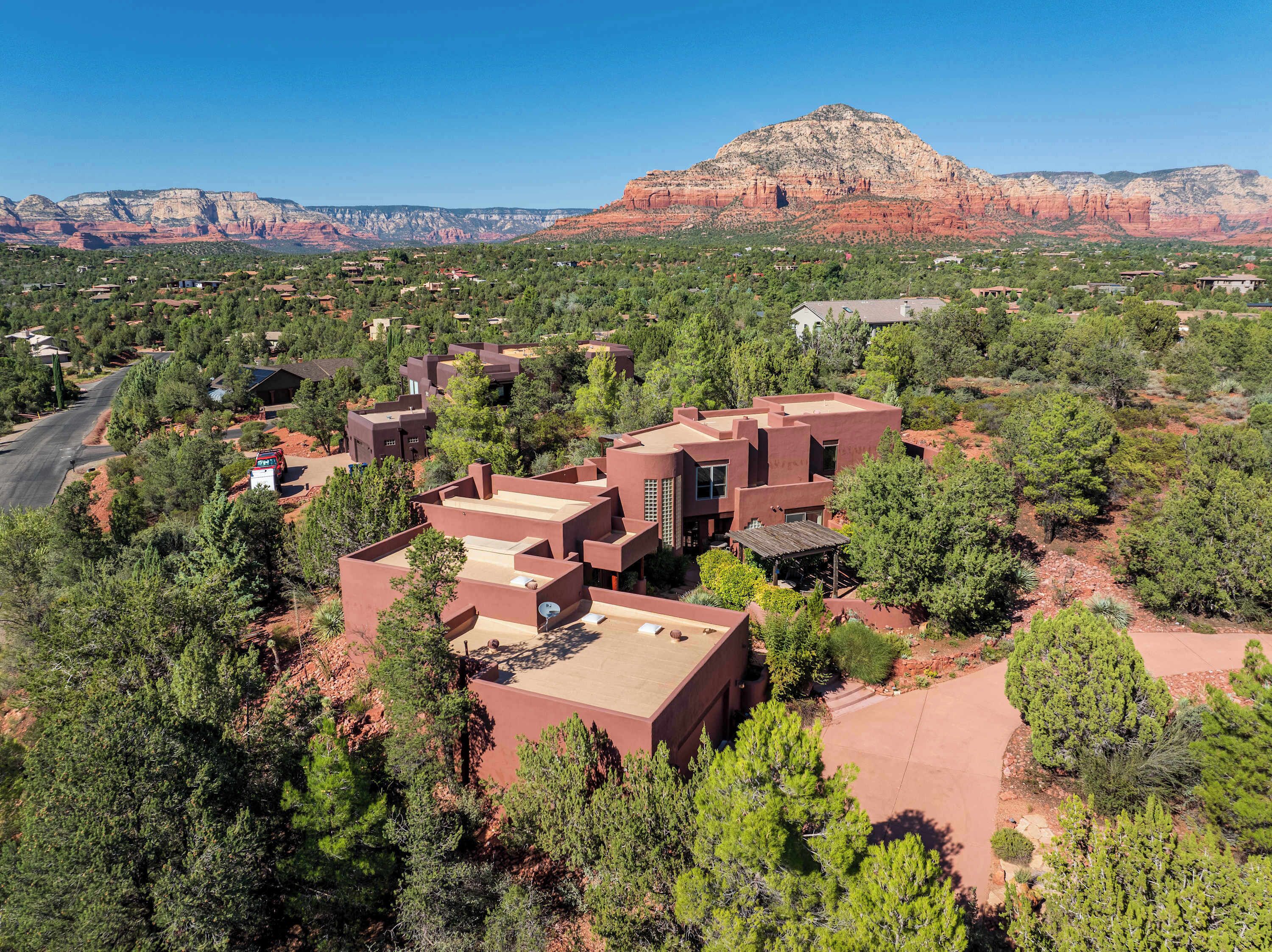 A residential area with houses featuring flat roofs and stucco exteriors, surrounded by dense green trees and a prominent red rock formation in the background.